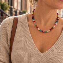 Load image into Gallery viewer, A woman wearing a colorful beaded necklace with a chunky knit beige V-neck sweater and white pants, carrying a brown shoulder bag in an outdoor urban setting.
